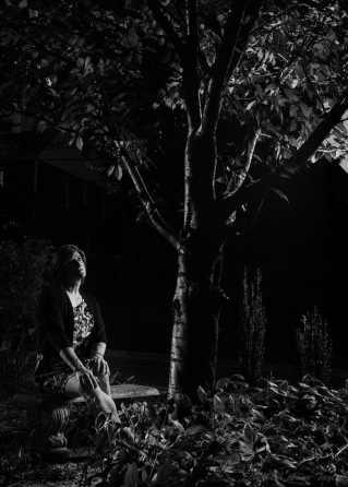 Black and white image of Allison sitting on a garden bench under a cherry tree. She and the tree are illuminated from opposite sides.
